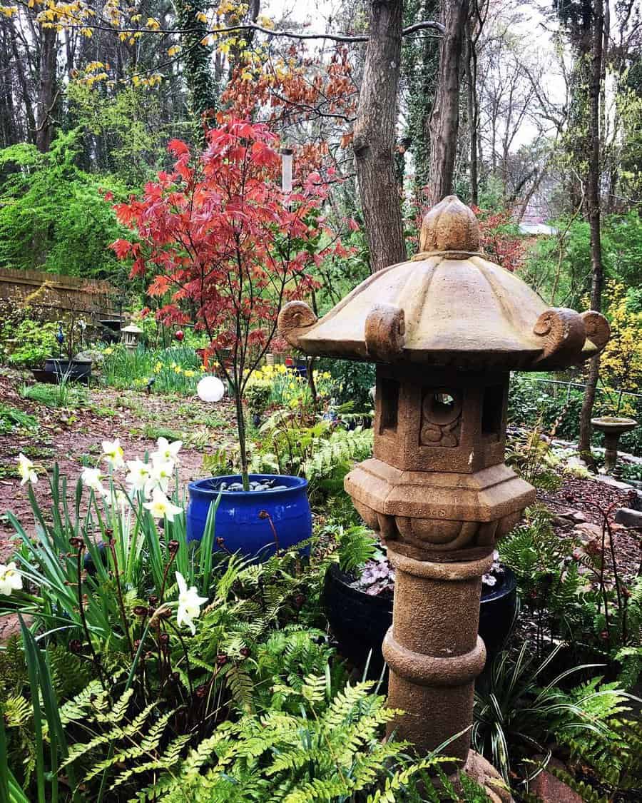 Serene garden scene with a stone lantern, red-leaved tree, blue pot, and white flowers surrounded by lush greenery and tall trees