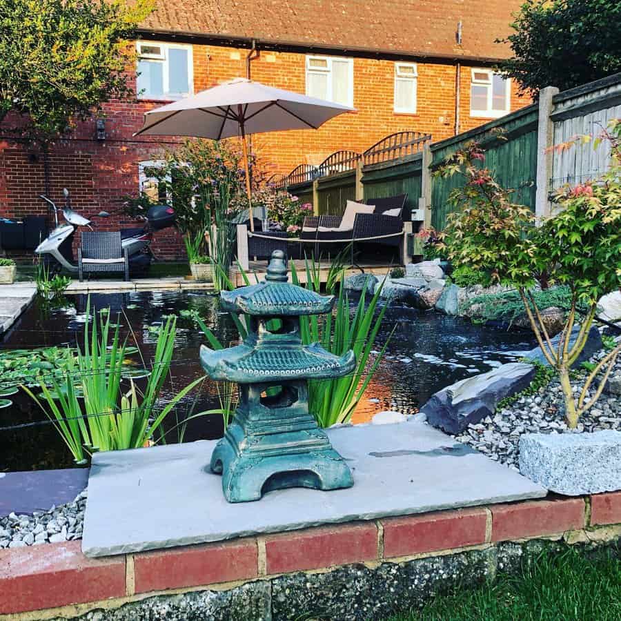 Garden scene featuring a pond, green stone lantern, plants, patio furniture, and a house in the background