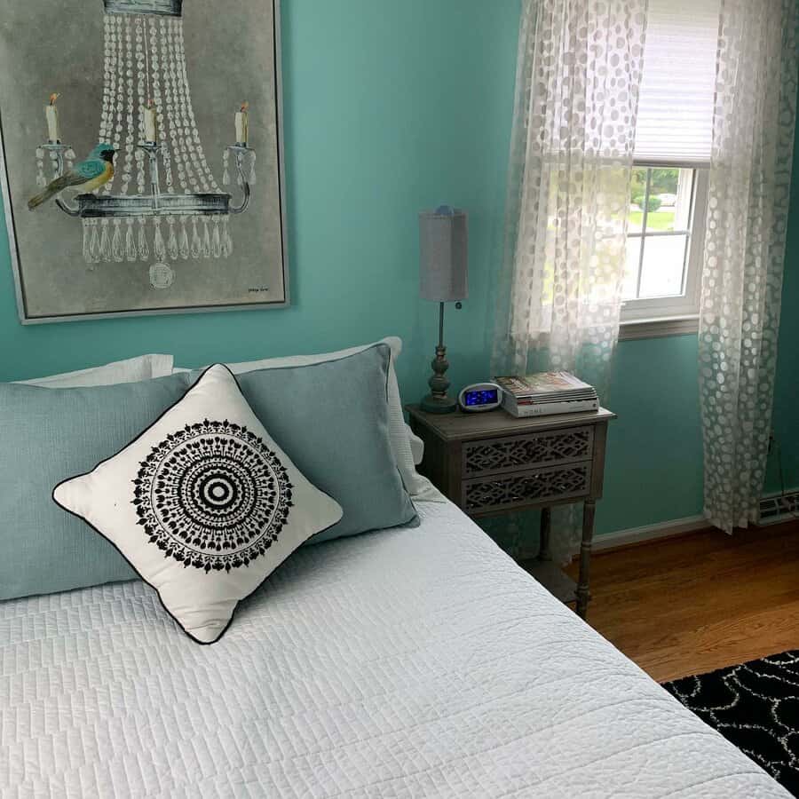 A cozy bedroom with mint walls, a bed with patterned pillows, a nightstand with books and an alarm clock, and a window with sheer curtains