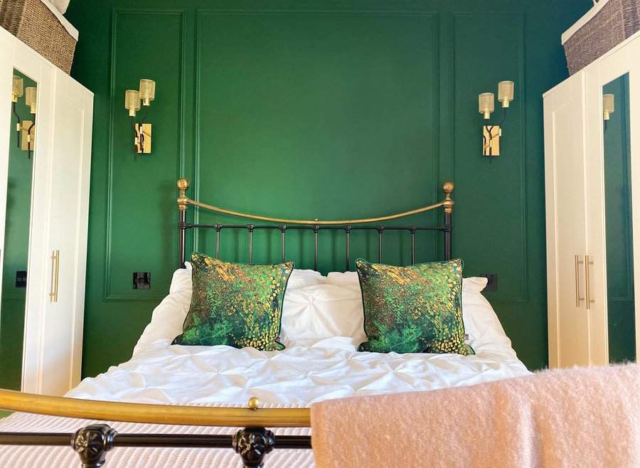 A cozy bedroom with a green accent wall, brass bedframe, white bedding, green patterned pillows, and beige lamps mounted on the wall