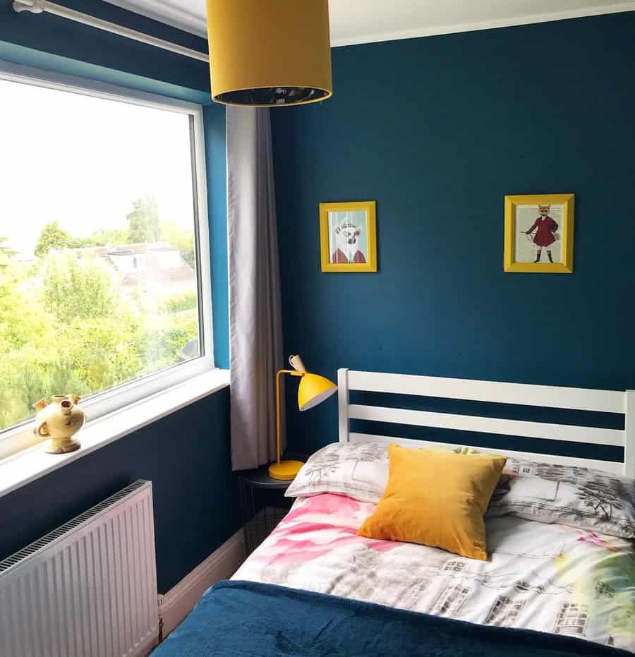 Cozy bedroom with blue walls, a white bed, yellow accents, and a large window with a view of greenery, two framed pictures are on the wall