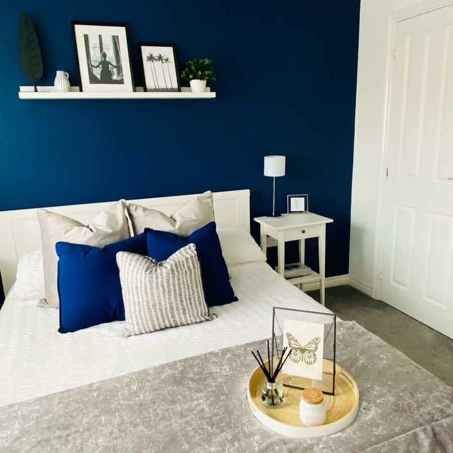 Stylish bedroom with white bed, blue and gray pillows, blue accent wall, art shelf, bedside table, and decorative tray on bed