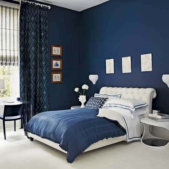 Stylish bedroom with blue walls, bedspread, and curtains; white bed with pillows, small side table, and artwork on the walls