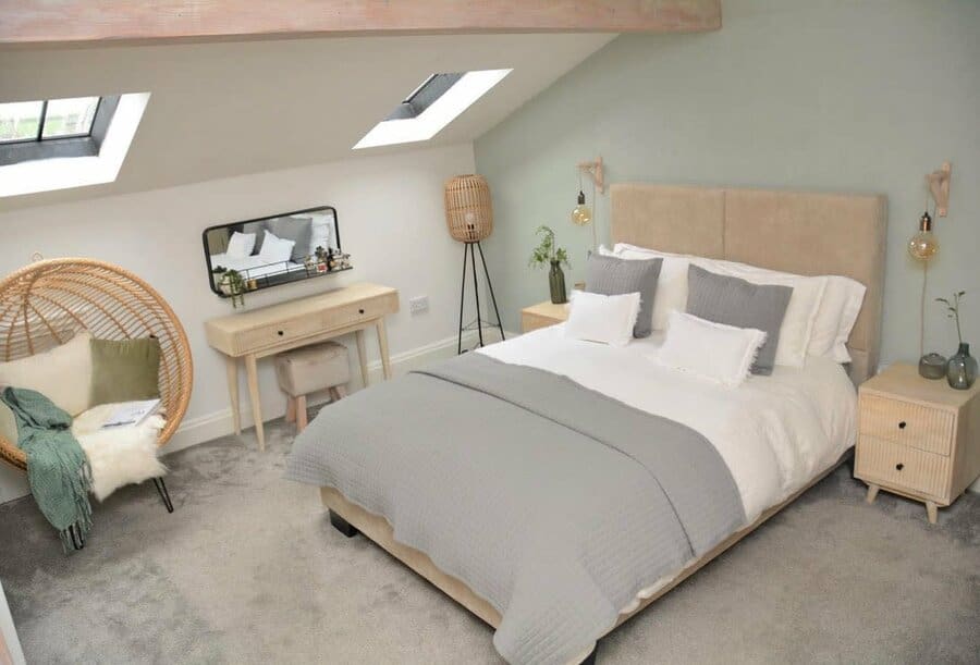 Cozy attic bedroom with skylights, a beige bed, gray accents, a wicker chair, and a vanity desk with a mirror