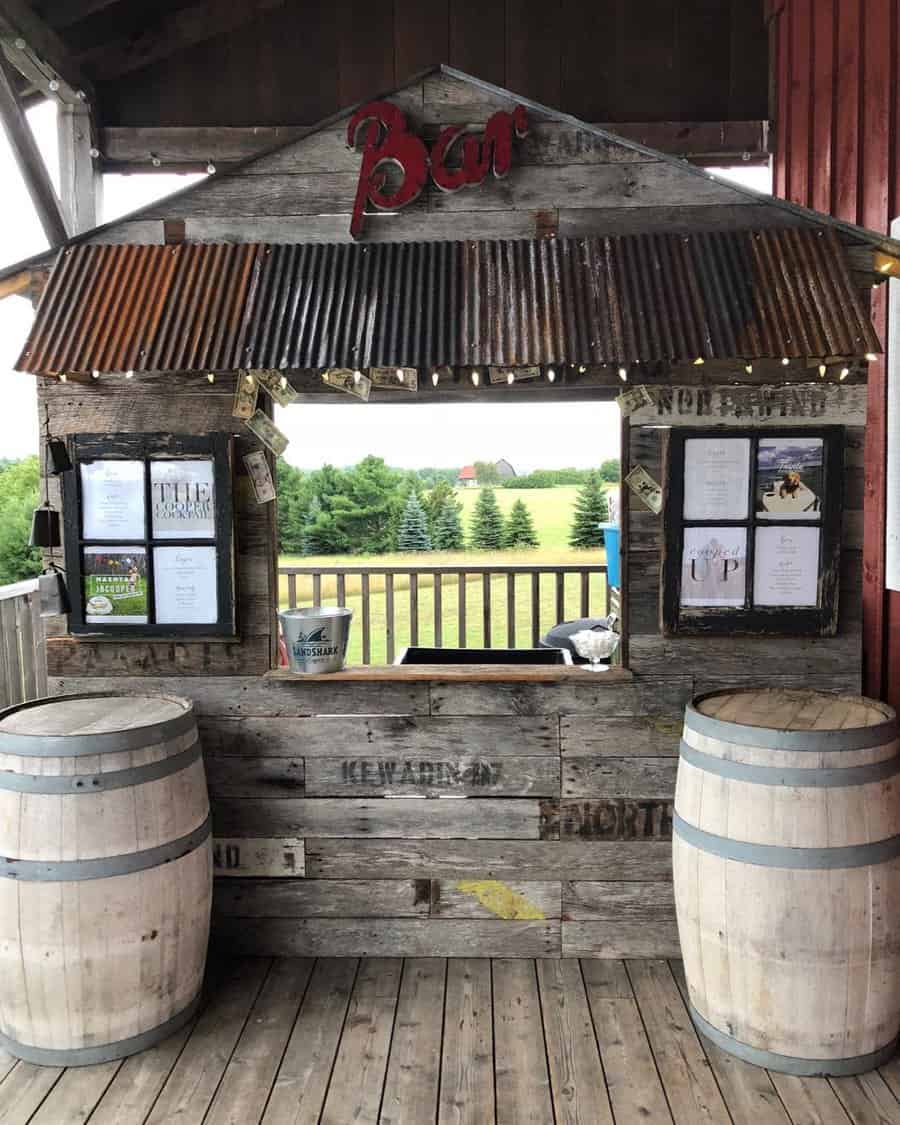 Rustic outdoor bar with corrugated metal roof, wooden barrels, and countryside view; menu displayed on the wooden wall