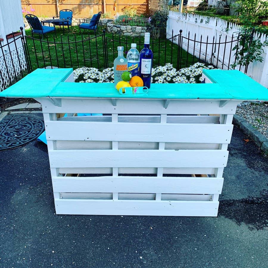 DIY bar made of white pallets with a turquoise top, featuring bottles and an orange on top in a backyard setting with flowers nearby