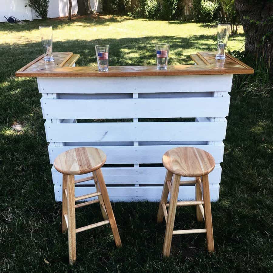 Outdoor bar made from white wooden pallets with a wooden top, featuring four glasses with flags, and two wooden stools on grass