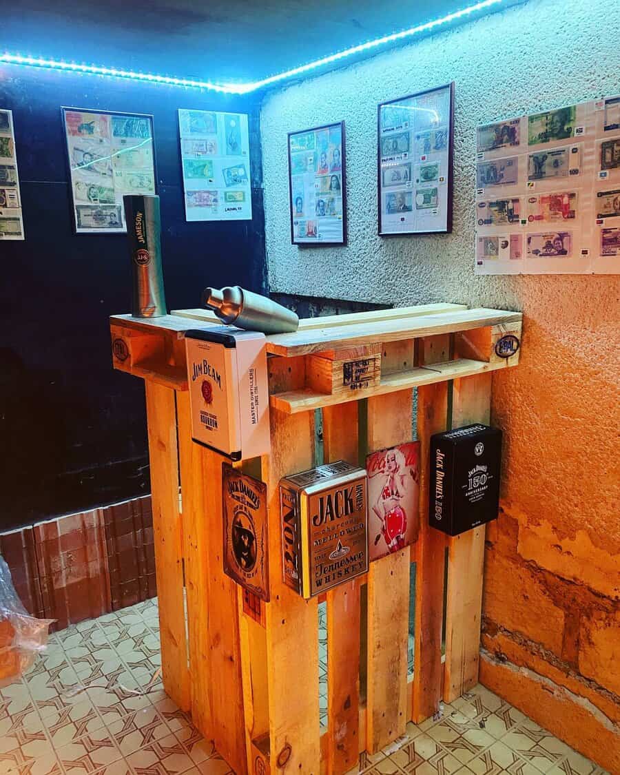 A rustic bar made of wooden pallets, decorated with liquor boxes and framed currency notes on the walls, under blue LED lighting