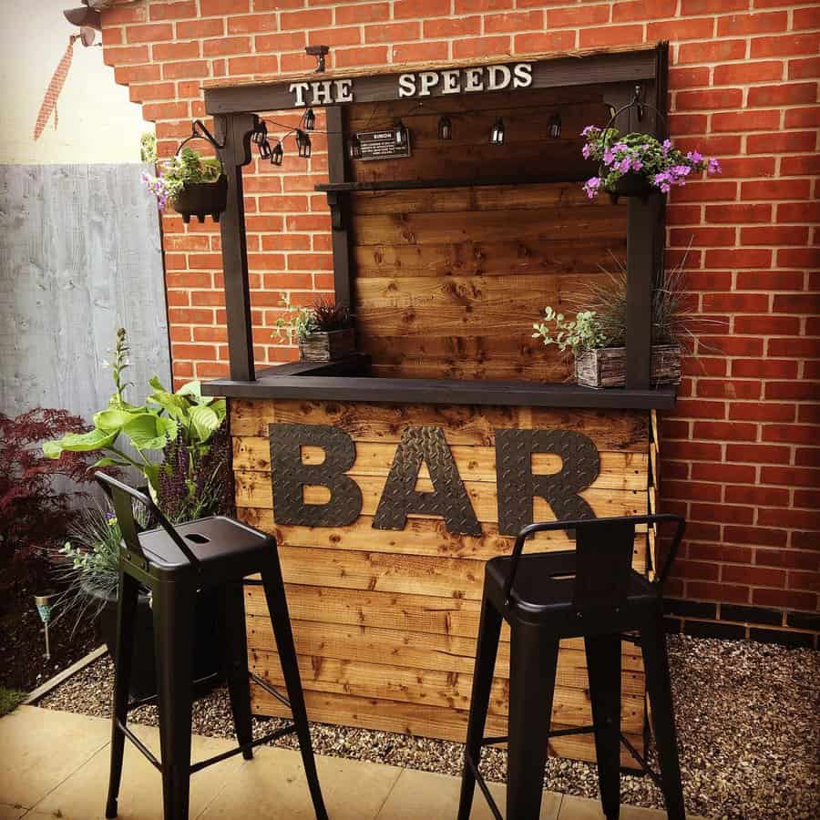 Rustic backyard bar with a wooden pallet design, black metal stools, and hanging plants, creating a stylish outdoor entertainment space