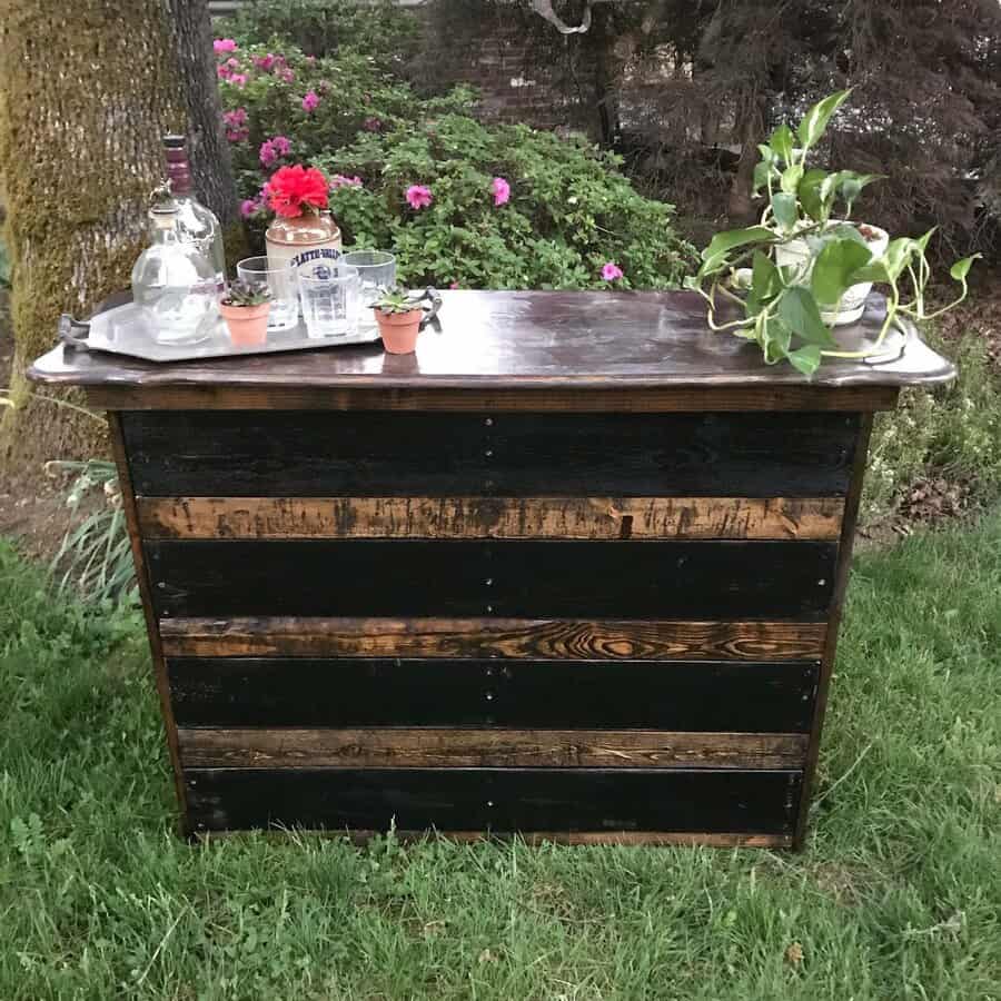 Outdoor bar with a wooden finish, topped with plants and bottles, set on grass with blooming flowers in the background