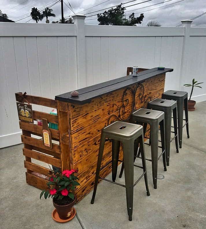 Outdoor wooden bar with three metal stools, plants, and a white fence in the background on a cloudy day