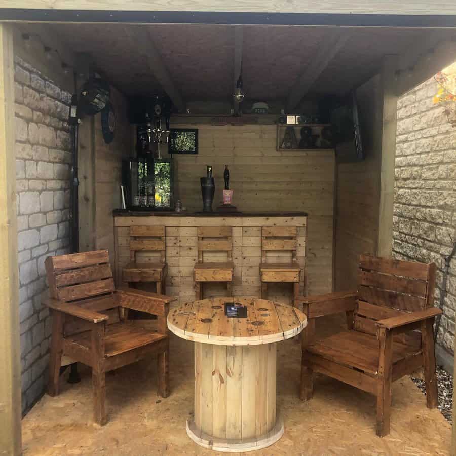 Rustic backyard bar with wooden pallet seating, a barrel-style table, and a cozy pub-like atmosphere, perfect for relaxing outdoors