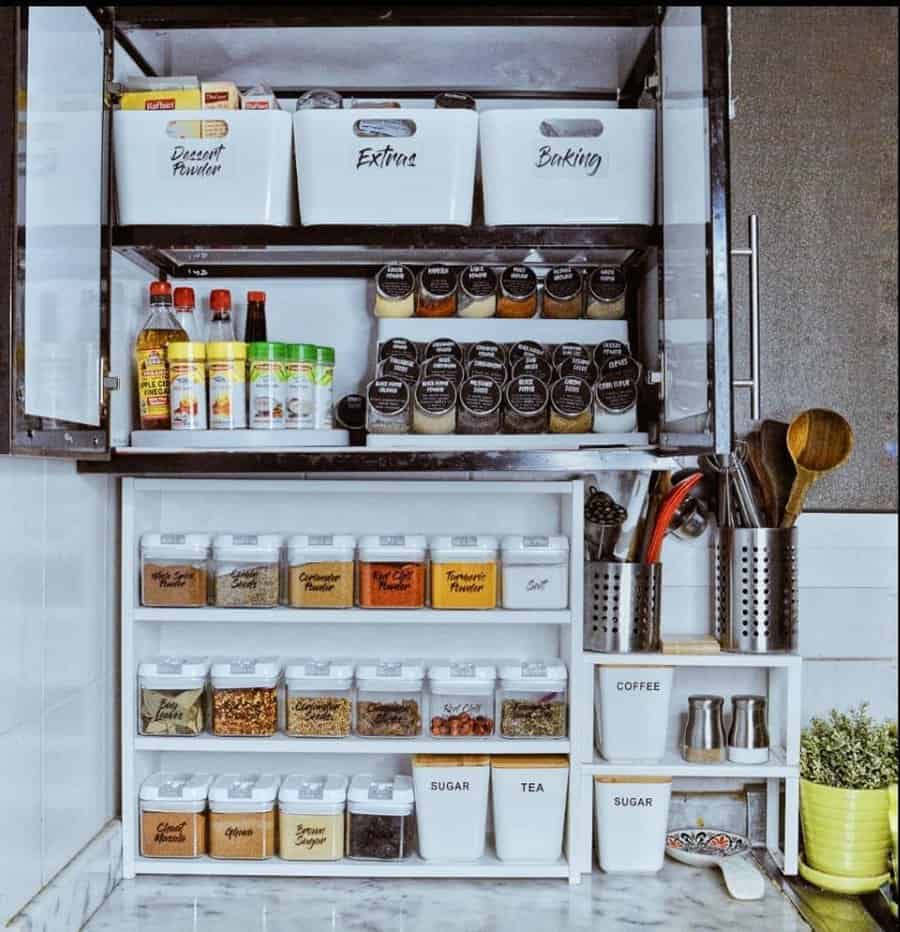 Organized kitchen cabinet with labeled containers for spices, baking supplies, and bottles, alongside utensils and mugs on a marble counter