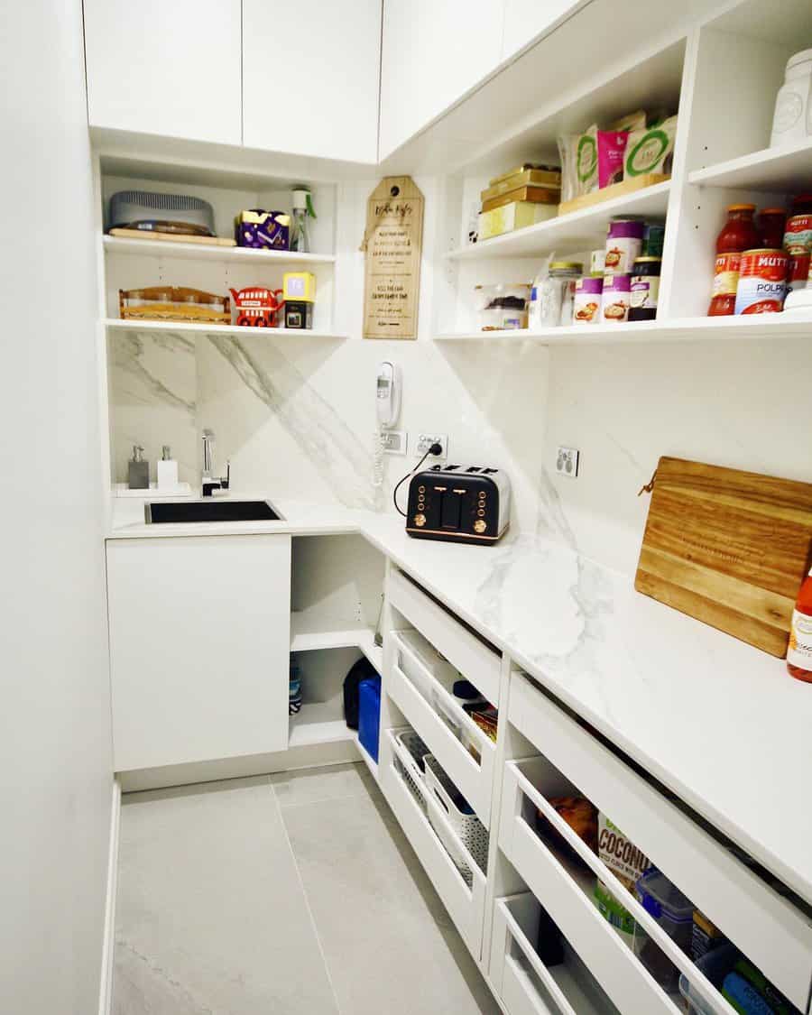 White pantry with open shelves, various food items, cutting board, small sink, toaster, and storage drawers along one wall