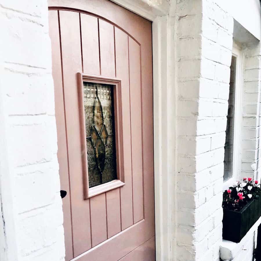 A pink door with a decorative glass inset, set in a white brick wall, with a window box of flowers beside it