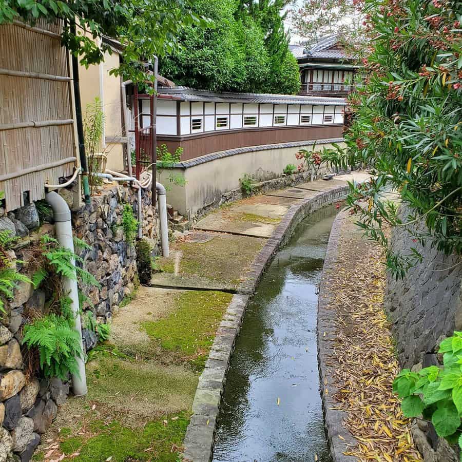 A narrow, curving canal with clear water runs between stone walls lined with greenery and traditional Japanese architecture