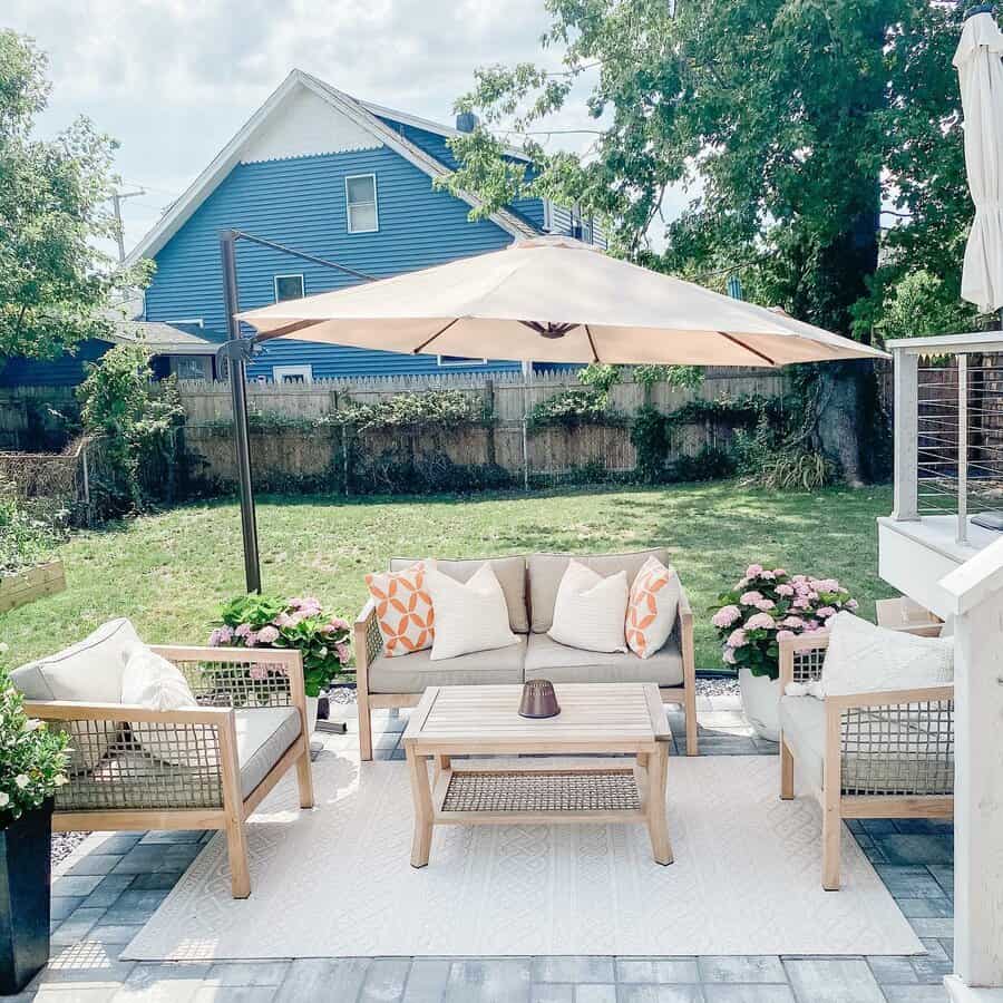 Outdoor patio with light wood furniture, beige cushions, and a cream umbrella. Blue house and garden with pink flowers in the background