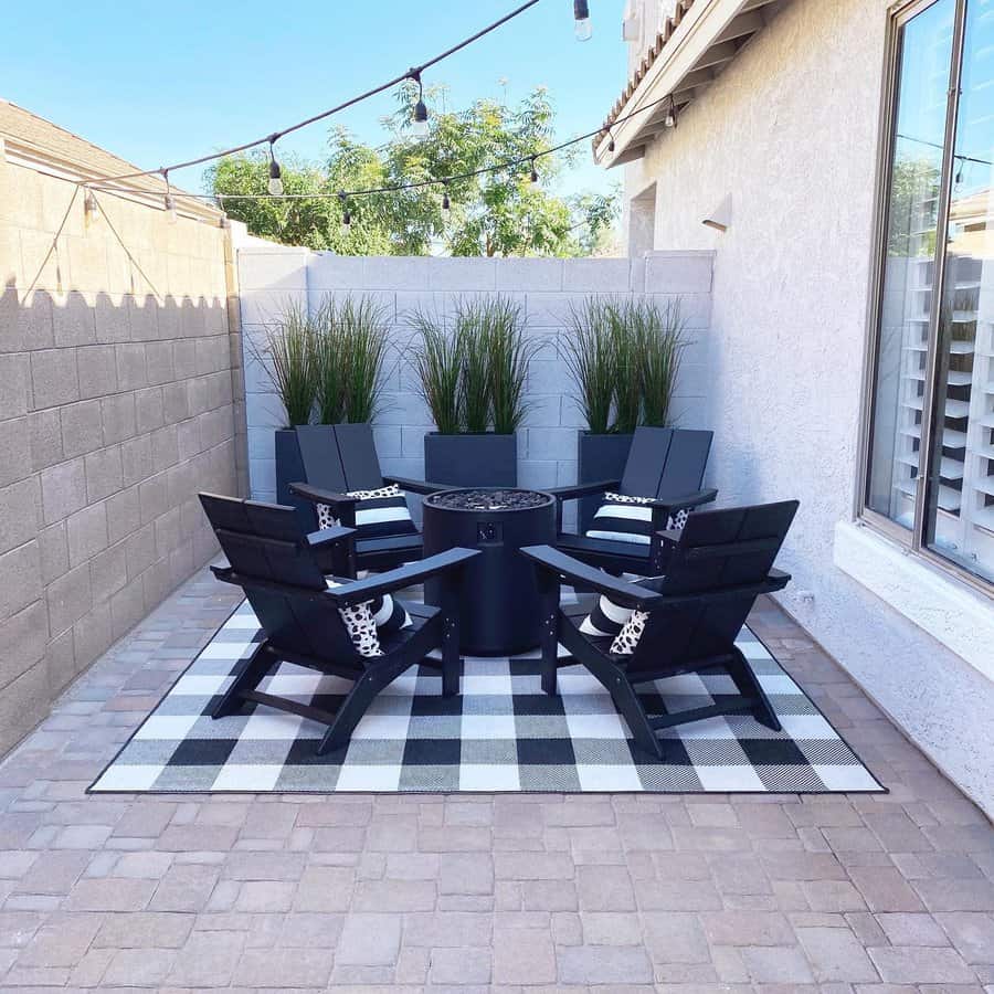 Outdoor patio with black chairs around a fire pit on a checkered rug. String lights are overhead, with tall plants against a wall