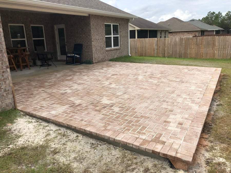 A backyard patio with light brown brick paving and a covered area attached to a brick house, next to a wooden fence