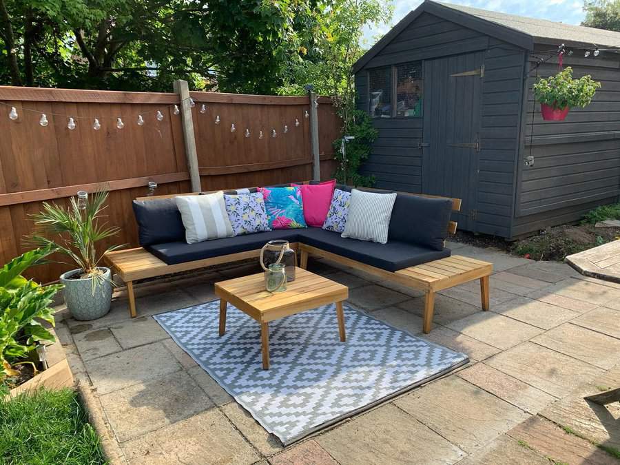 Cozy backyard patio with stone pavers, a wooden L-shaped bench with colorful cushions, a stylish rug, and string lights for a relaxing outdoor vibe