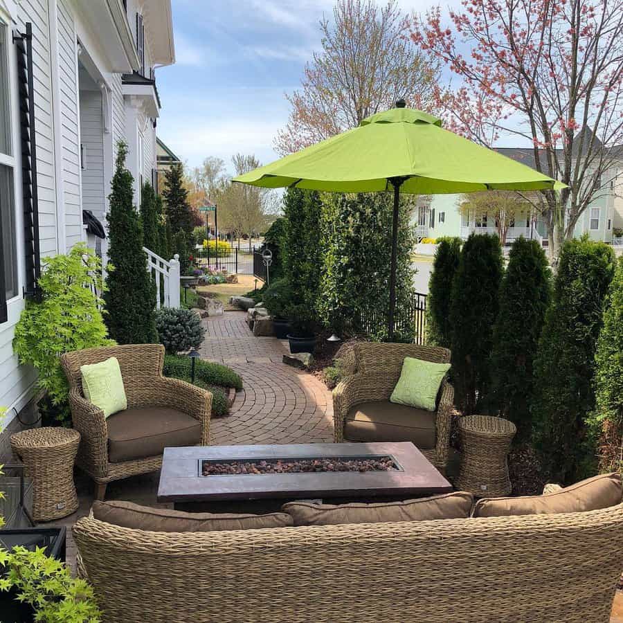 Elegant backyard patio with curved brick pavers, wicker seating, a fire table, lush greenery, and a bright green umbrella for a cozy outdoor retreat