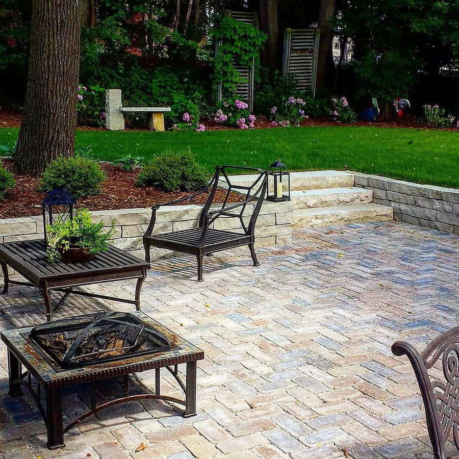 Charming backyard patio with herringbone brick pavers, wrought iron seating, a fire pit, and lush greenery for a cozy and elegant outdoor retreat