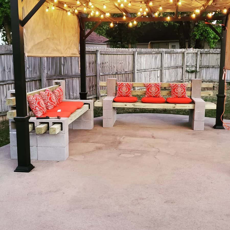 DIY outdoor lounge area with cinder block benches, wooden seating, vibrant red cushions, and string lights under a pergola, creating a cozy backyard retreat