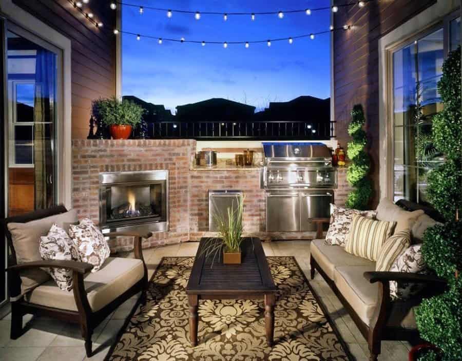 Cozy patio with sofas, coffee table, fireplace, and grill; string lights above and a patterned rug on the floor, plants on either side