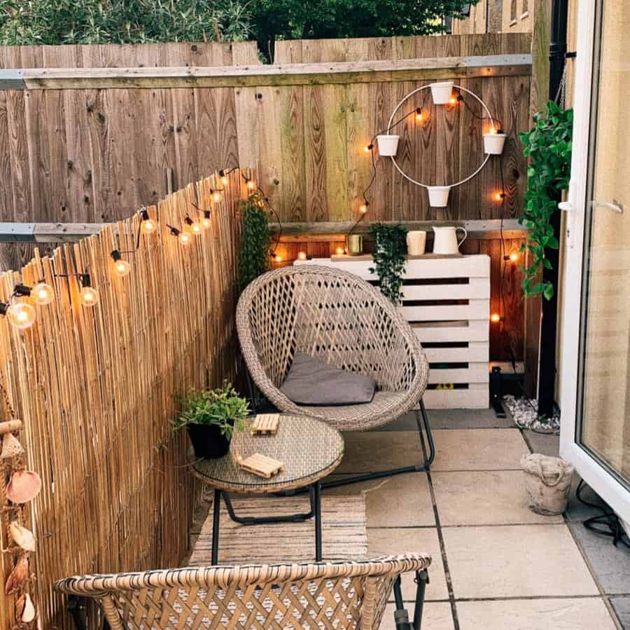 Cozy patio with wicker chairs, a glass table, string lights, and potted plants, surrounded by a wooden fence