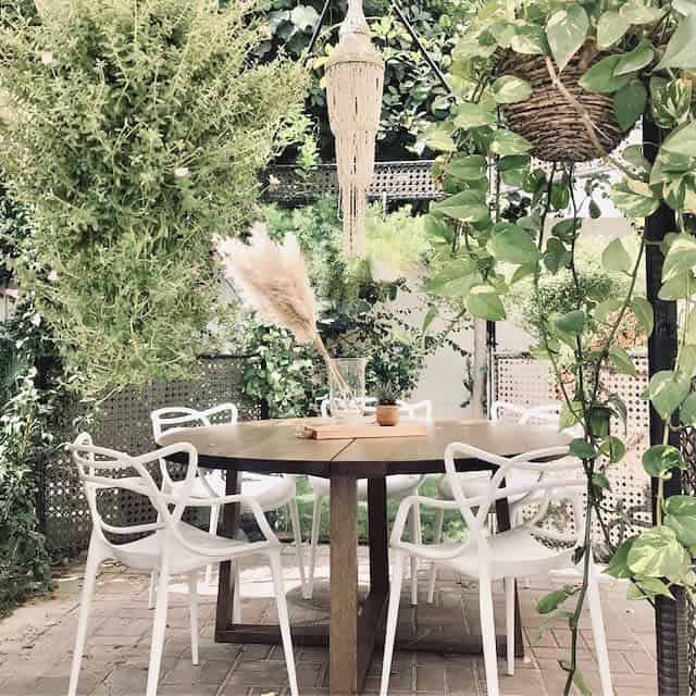 Outdoor dining area with a wooden table, white chairs, hanging plants, and greenery, pampas grass in a vase on the table