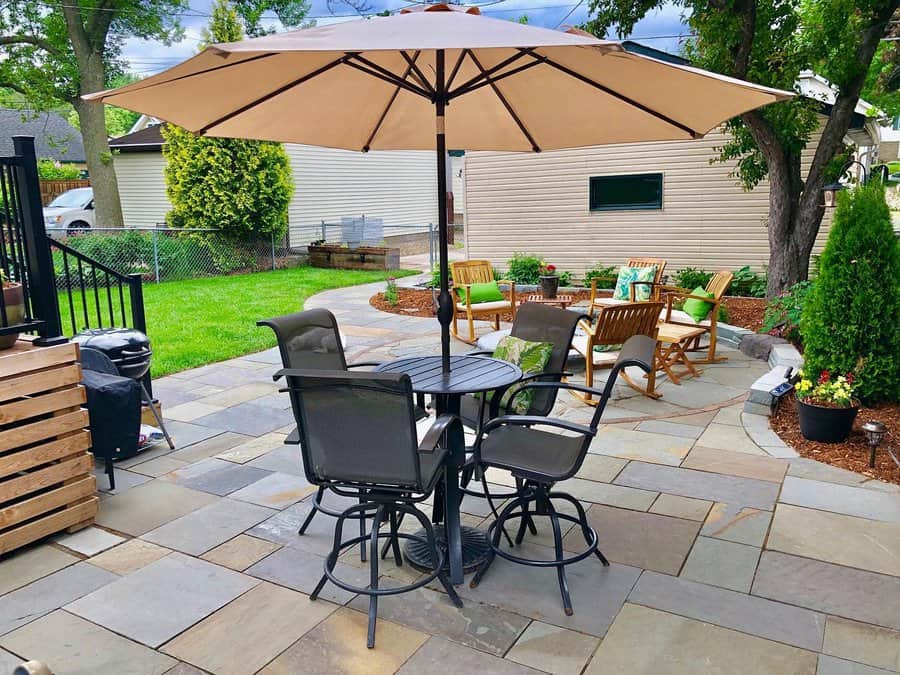 Backyard patio with stone tiles, a table, chairs under a large umbrella, and additional seating surrounded by greenery