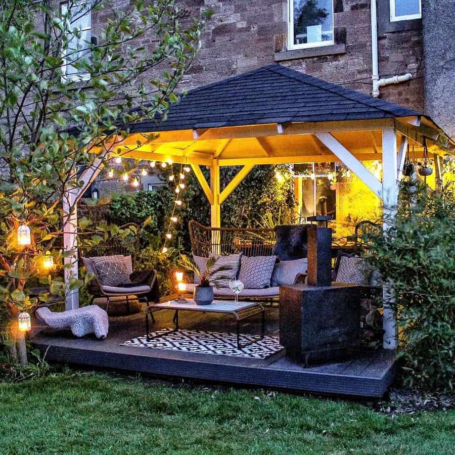 Cozy outdoor gazebo with string lights, cushioned chairs, and a table on a patterned rug; lush greenery surrounds the setting