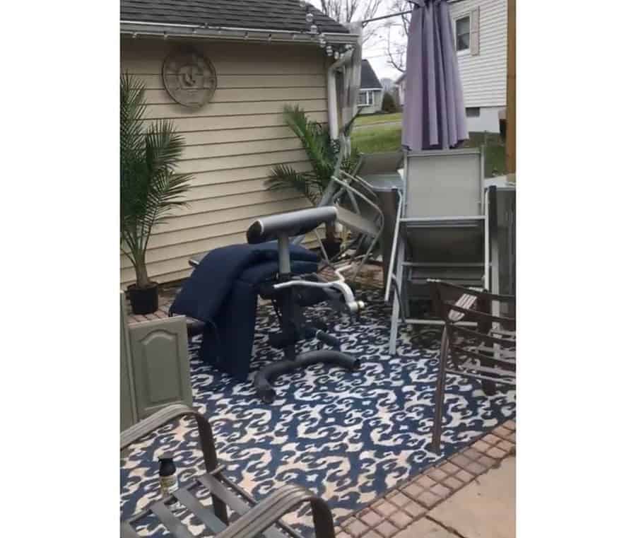 Backyard with overturned patio chairs, umbrella, and furniture, blue patterned rug on the ground, brick border, and two potted plants