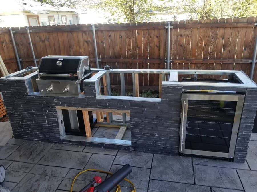 Outdoor kitchen under construction with a built-in grill on the left and a mini fridge on the right, against a wooden fence