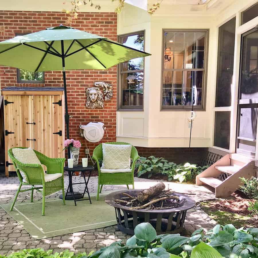 Cozy patio with green wicker chairs, umbrella, firepit, and flowers; brick wall backdrop and a wooden cabinet on the side