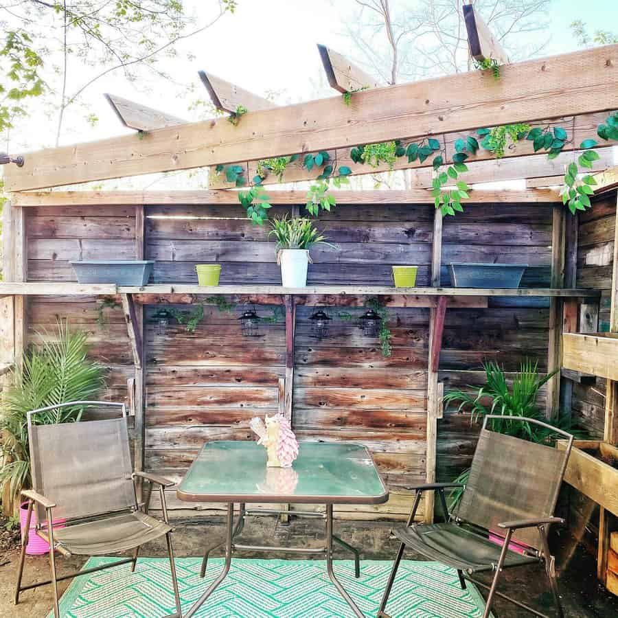 Rustic patio with a wooden pergola, greenery, and a cozy seating area featuring a glass-top table and metal chairs on a patterned rug