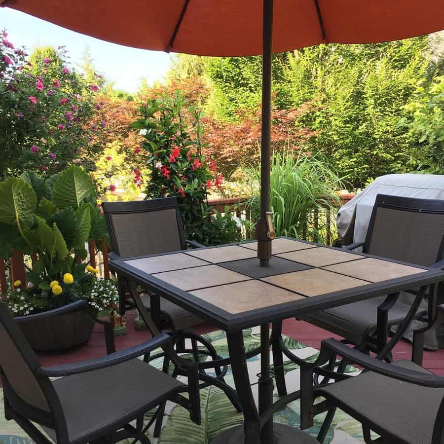 Patio table with four chairs under an orange umbrella, surrounded by lush green plants and colorful flowers