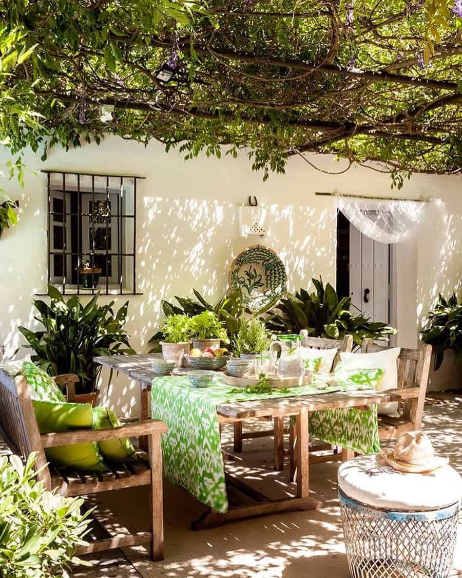 Charming Mediterranean-style patio with a wooden dining set, lush greenery, and natural shade from vine-covered branches overhead