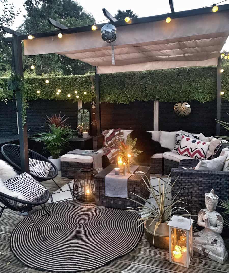 Boho-style patio with a pergola, string lights, wicker seating, cozy cushions, candles, a black accent wall, and lush greenery