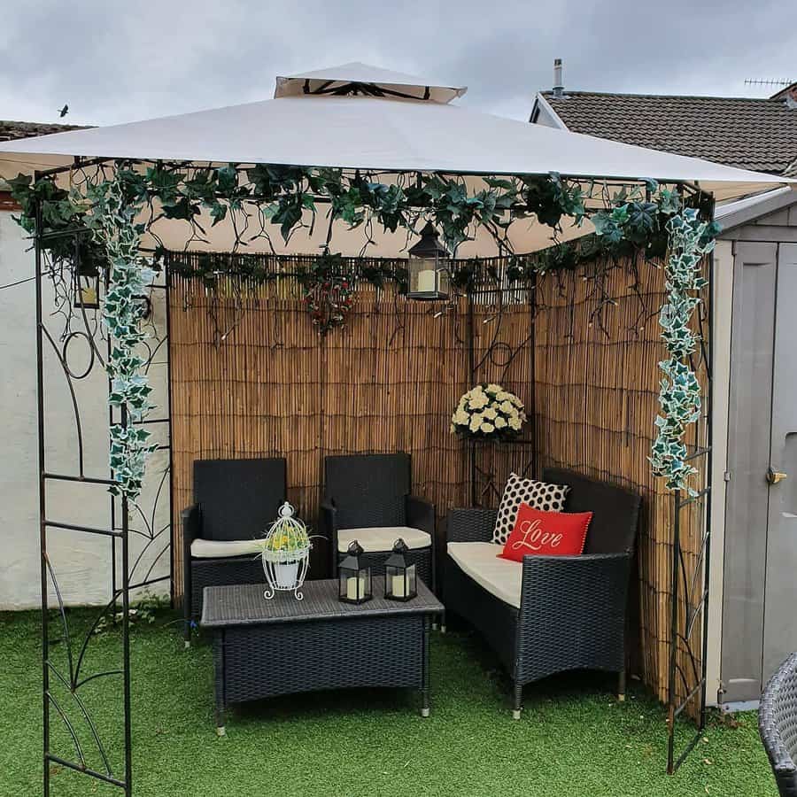 Cozy gazebo with wicker furniture, bamboo privacy screen, faux ivy decor, lanterns, and a 'Love' pillow on artificial grass