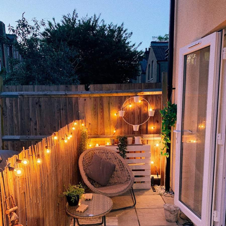 Cozy patio with warm string lights, wicker chair, small table, potted plants, and a circular wall planter on a wooden fence