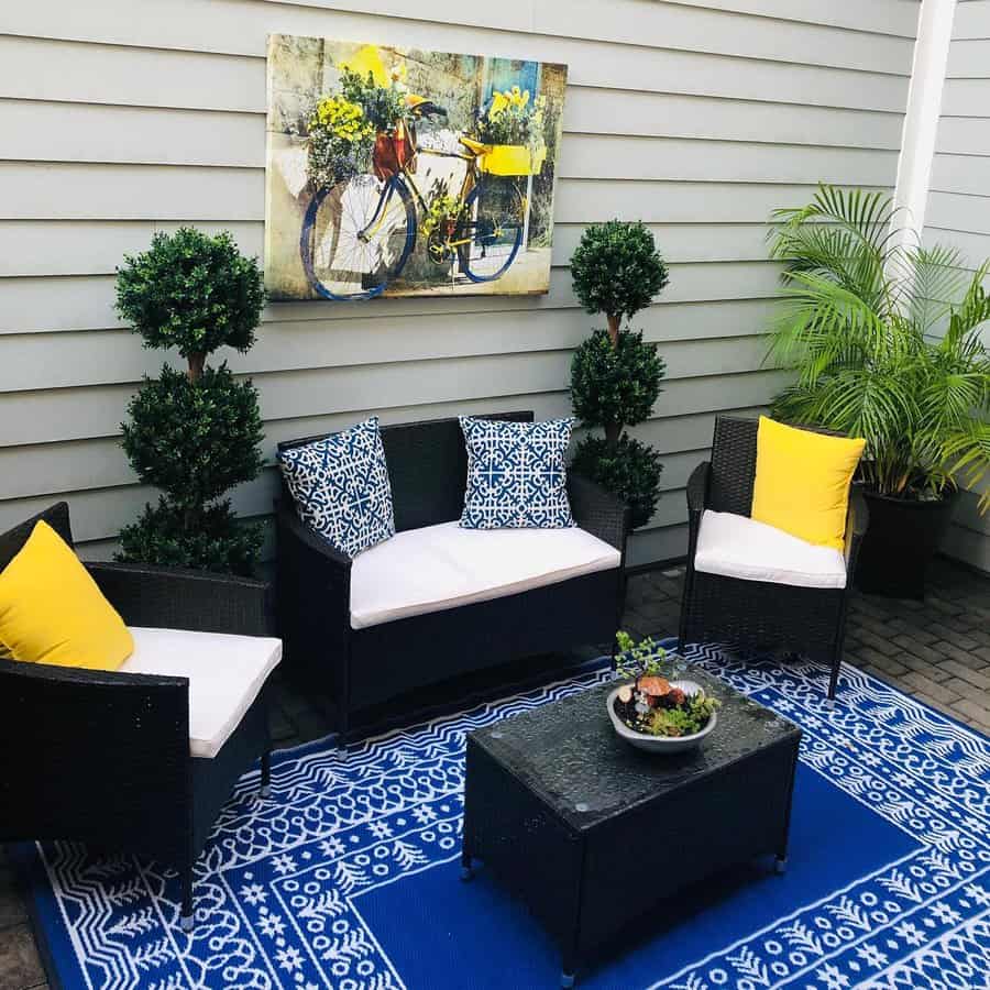 Outdoor patio with black wicker furniture, blue and white cushions, yellow accents, a blue patterned rug, plants, and bike art on the wall