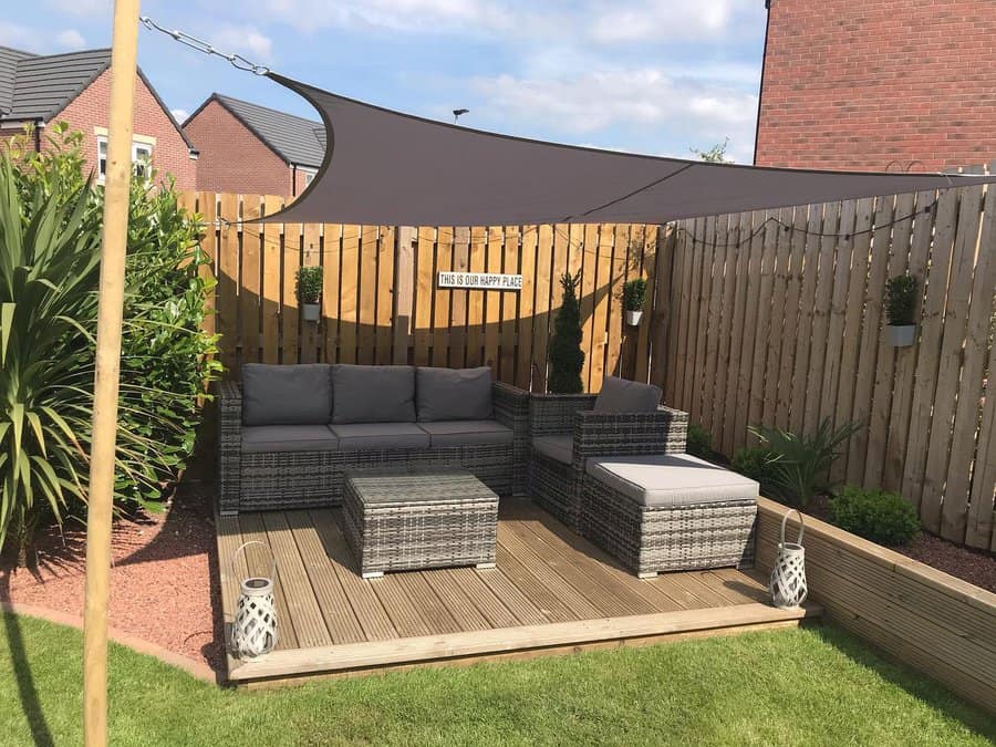 Cozy backyard patio with a dark shade sail, rattan furniture, wooden decking, and potted plants, creating a relaxing outdoor space