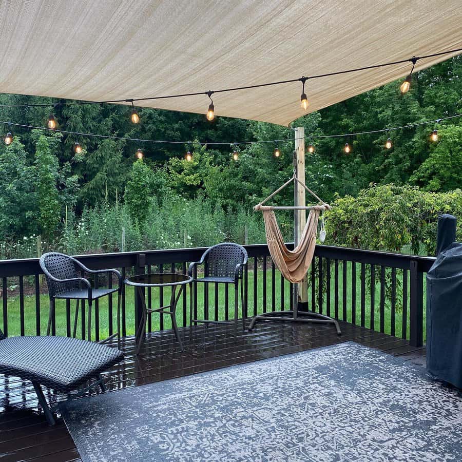 Cozy backyard deck with a beige shade sail, string lights, a hammock chair, and black wicker furniture, surrounded by lush greenery