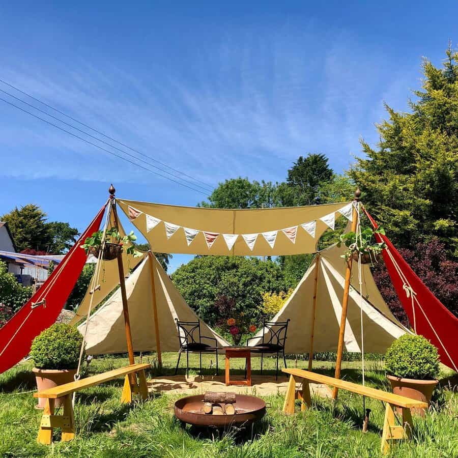 Charming outdoor seating area with shade sails, a fire pit, wooden benches, and decorative bunting, set in a lush green garden