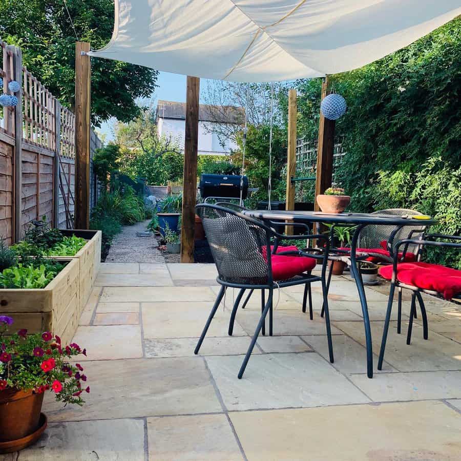 Cozy backyard patio with a white shade sail, outdoor dining set with red cushions, potted plants, and a barbecue grill