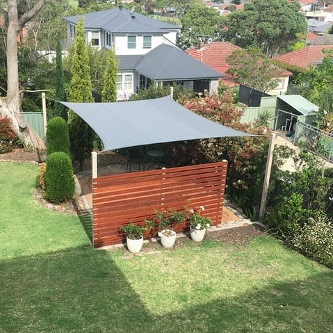 Backyard patio with a grey shade sail, wooden privacy screen, and potted plants, creating a shaded outdoor seating area