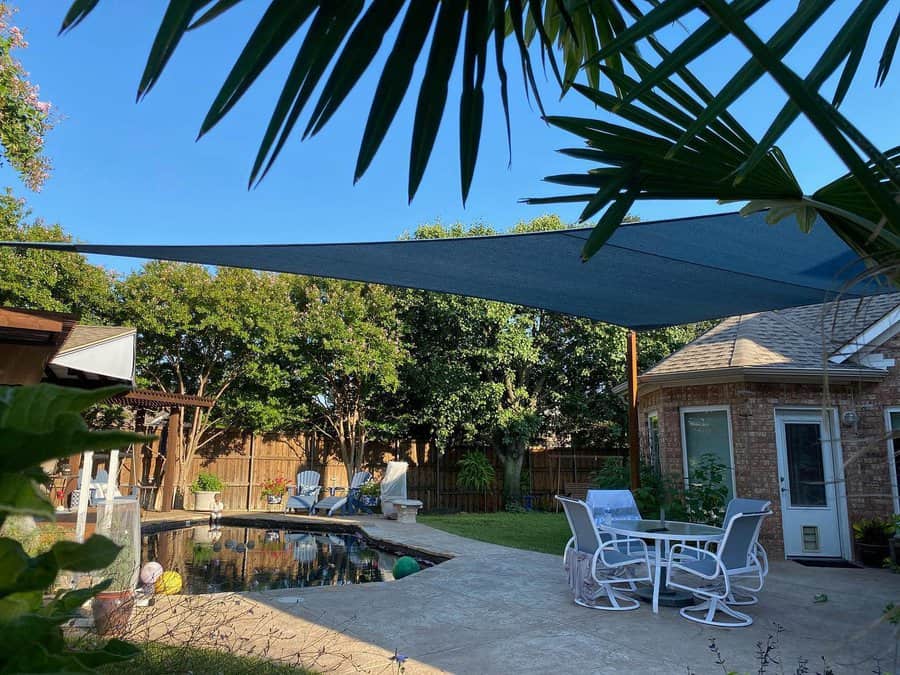 Backyard poolside patio with a blue shade sail, white outdoor dining set, lounge chairs, and lush greenery