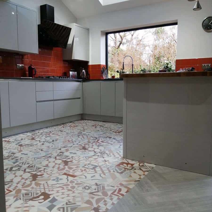 Modern kitchen with gray cabinets, red backsplash, patterned floor tiles, skylight, and large window overlooking trees