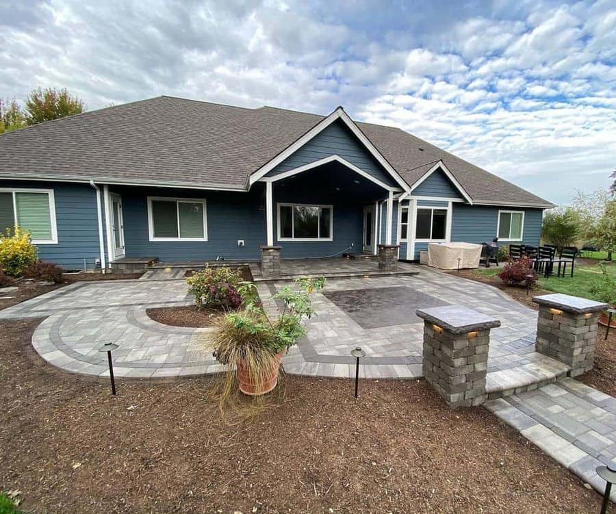 Large backyard patio with interlocking stone pavers, decorative pillars, and landscaping around a modern blue house